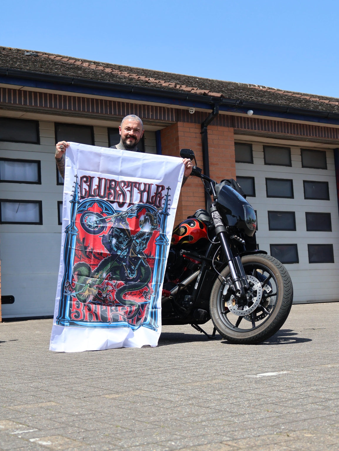 A rider holding a Clubstyle Britain banner beside a custom motorcycle.